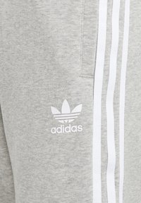 Pilki medvilniniai kelnės su baltomis šoninėmis juostelėmis ir siuvinėtu baltu Adidas logotipu, kuriame pavaizduoti trys lapai. Minkšta tekstūra su kasdiene dizainu.