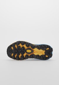Semelle d'une chaussure de randonnée bleue et jaune avec des crampons en caoutchouc noir conçus pour l'adhérence sur un terrain accidenté, affichée sur un fond blanc.