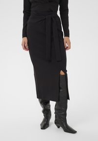 Melna midi svārki ar sānu šķēlumu un sasieto auduma jostu. Gluda tekstūra, pieguļošs dizains, apvienoti ar melniem augstiem ādas zābakiem līdz ceļgaliem.