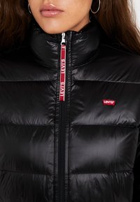 Žena v černé prošívané bundě Levi's s červenou nášivkou s logem a červeným táhlem zipu, které nese značku Levi's.