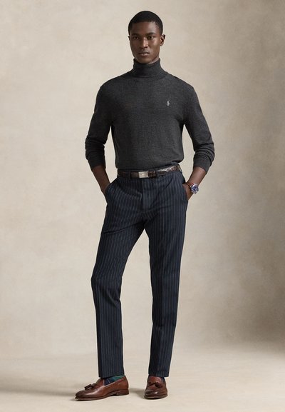 Young man wearing a dark gray turtleneck, navy pinstripe trousers, brown leather belt and loafers, standing with hands in pockets.