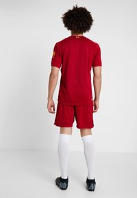 Röd fotbollströja med korta ärmar, matchande röda shorts och vita knähöga strumpor. Svarta fotbollsskor med grå accenter. Platt bakvy.