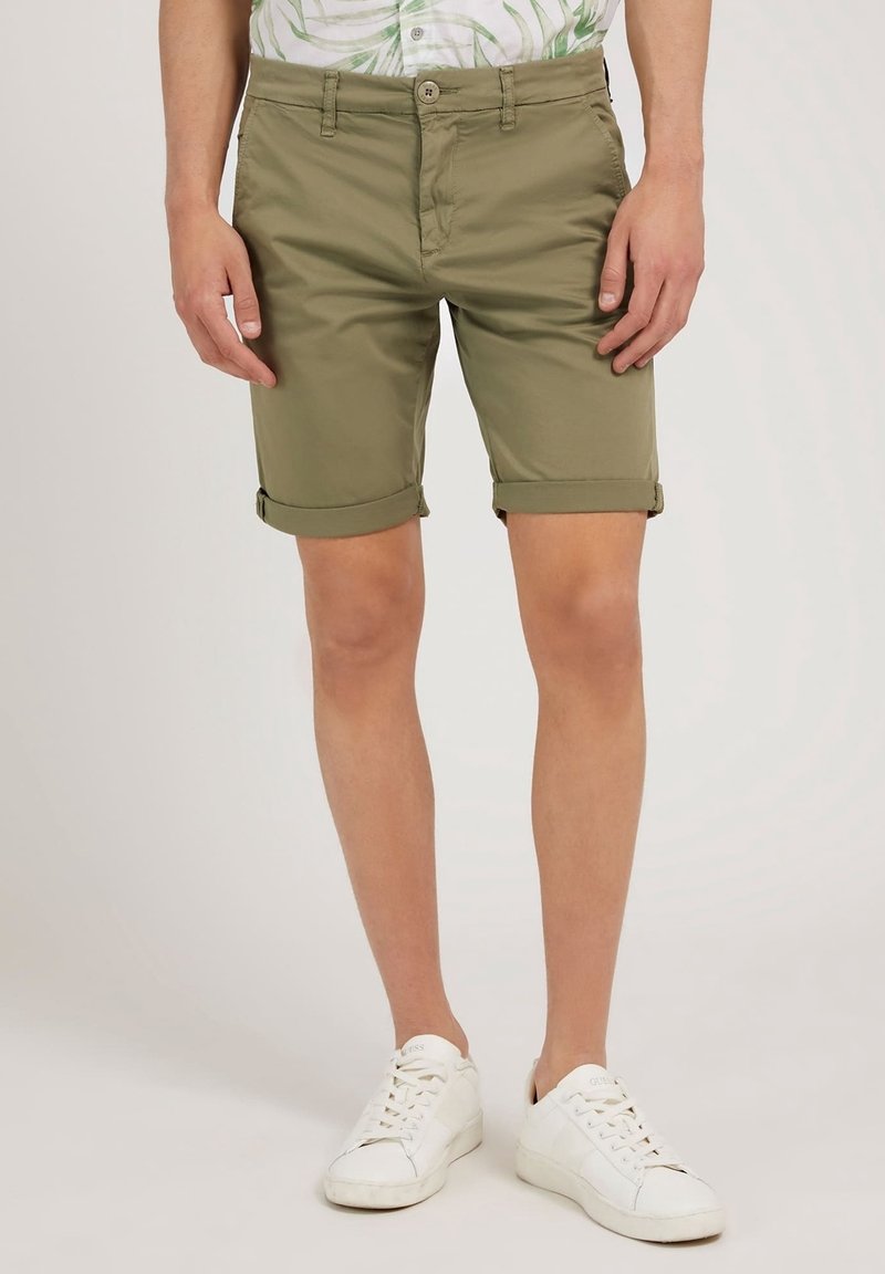 Pantalones cortos de algodón verde oliva con dobladillo enrollado, cierre de botón en el frente y presillas para cinturón, combinados con zapatillas blancas.
