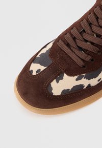 Adidași din suede maro cu panouri din țesătură cu imprimeu de vacă, având șireturi rotunde și talpă din cauciuc. Textură netedă cu un element de design contrastant.