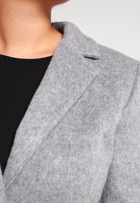 Blazer grigio in lana con rever a scollo, texture in feltro e chiusura a un bottone, indossato sopra un top nero. Visione ravvicinata del colletto e del tessuto.