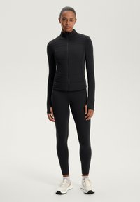 Donna in piedi che indossa una giacca a zip nera trapuntata e leggings neri con scarpe da corsa beige, su uno sfondo chiaro e semplice.
