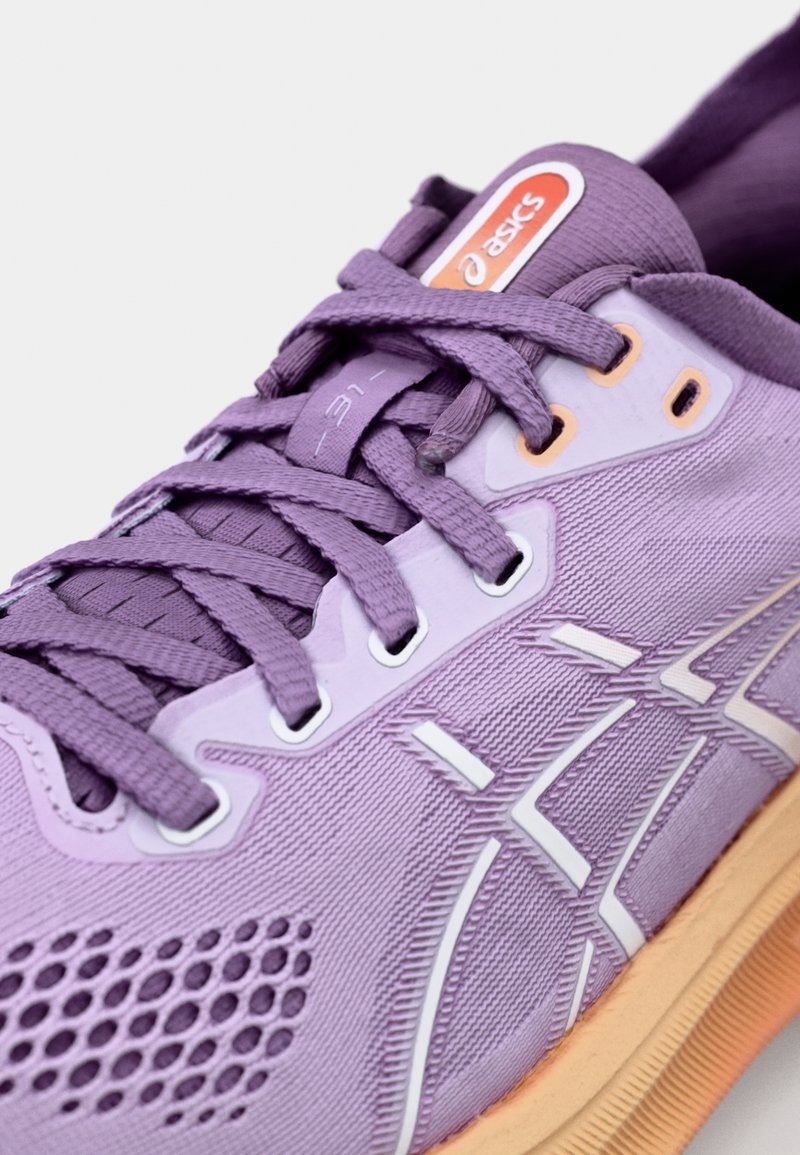 Zapatilla deportiva morada con un upper texturizado, acentos en blanco y un panel de malla visible para transpirabilidad. Incluye detalles de marca en naranja y blanco.