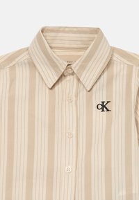Béžová a krémová vertikálne pruhovaná košeľa na gombíky s golierom a čiernym vyšívaným logom "cK" na ľavej hrudi.