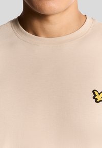 T-shirt in cotone beige con scollatura rotonda e maniche corte. Presenta un logo di un uccello ricamato giallo nella parte sinistra del petto.