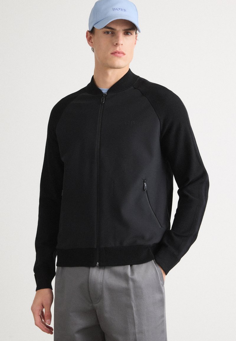 Svart zip-up jakke med strikket tekstur, ribbede mansjetter og kant. Har en diskret logo og en sidelomme med glidelås. Kombinert med en lyseblå caps.