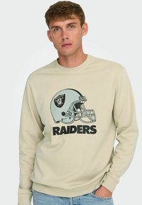 Beige Sweatshirt mit Rundhalsausschnitt, mit einem Grafikdesign eines Footballhelms und dem Wort "RAIDERS" in fetten schwarzen Buchstaben.