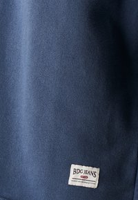 Tamnoplava tkanina s mekom teksturom, s malom bijelom etiketom na kojoj piše "BDG JEANS" s crvenom ivicom i detaljima o proizvodnji.