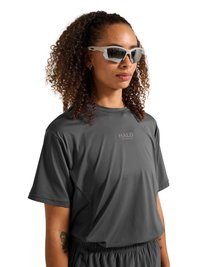 Woman with curly hair wearing reflective white sports sunglasses and a dark gray HALO logo t-shirt, looking forward.