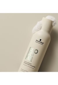 Flacone di shampoo bianco con testo argentato, etichettato "BLONDME Bond Repair Nourishing Shampoo", ricoperto da schiuma bianca, su uno sfondo beige chiaro.