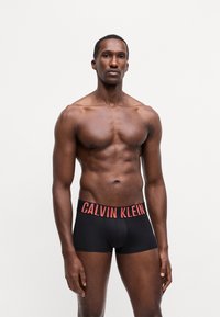 Črne bokserice z rdečim pasom z napisom "CALVIN KLEIN", gladka tkanina, prilagojen dizajn, prikaz modela z atletskim telesom.
