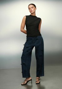 Naine seljas ilma põhjuseta must sodi, laiad tume teksad, millel on väikesed paelad, ja mustad rihmadega kontsakingad, seismas neutraalse taustaga.