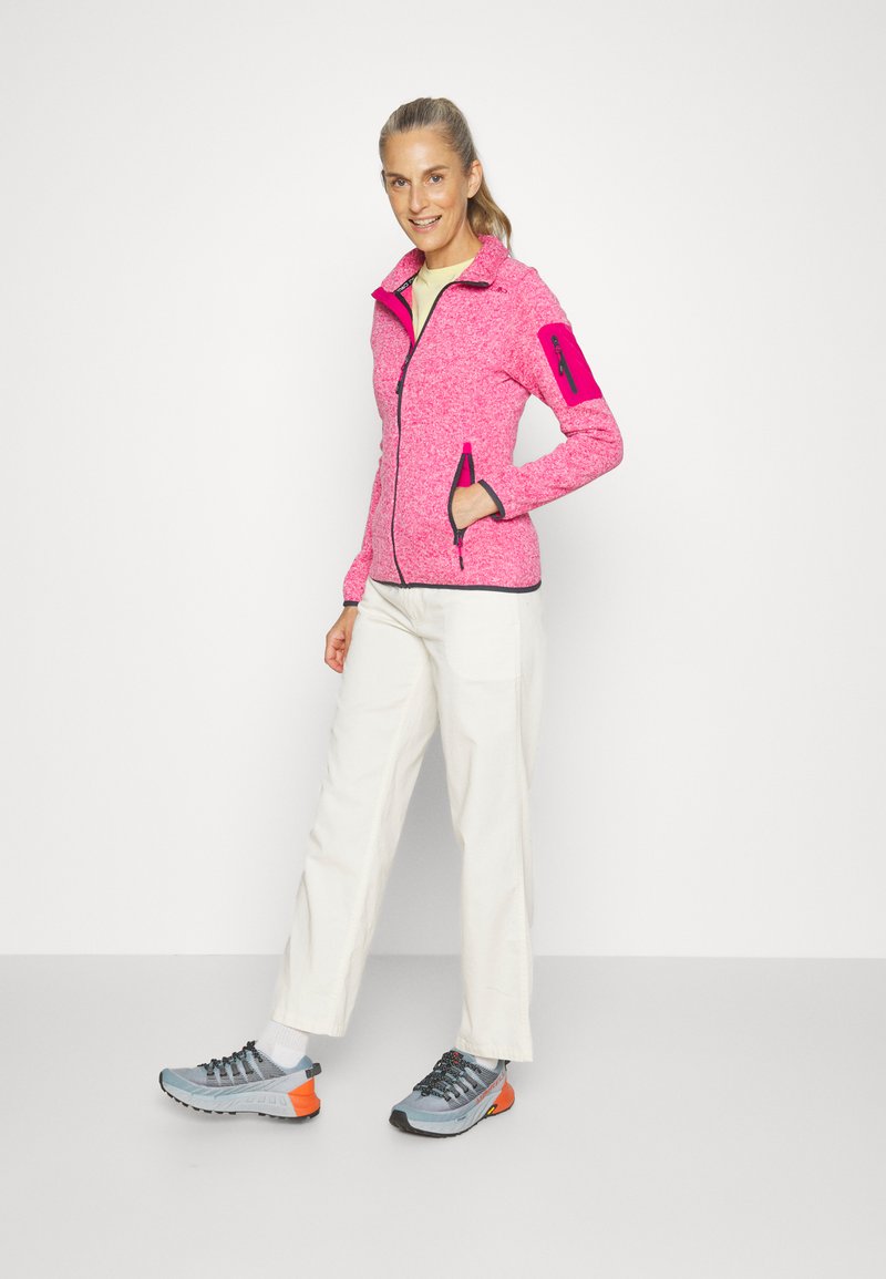 Giacca in fleece rosa con colletto alto, tasche con zip e zip nere a contrasto. Abbinata a pantaloni beige e scarpe grigio-arancio.