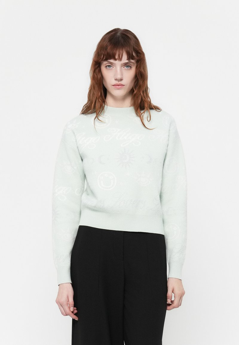 Mintgroene trui met een ronde hals, versierd met lichte symbolen en tekst. Zachte textuur, nauwsluitend in de taille, gecombineerd met zwarte broeken.
