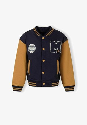 Veste universitaire marine et moutarde à fermeture par boutons, col et poignets rayés, écusson avec "M" sur la poitrine, et emblème "Game Day" sur le devant.