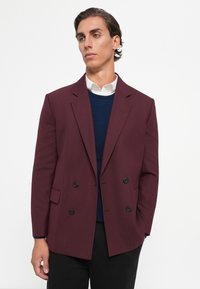 Bordeaux dobbelbrystet blazer med sorte knapper, spidse klapper og en glat overflade. Båret over en marineblå sweater og en hvid skjorte.