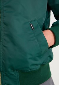 Zelena bomber jakna napravljena od glatkog, sjajnog materijala. Sadrži rebraste manžetne i porub, zatvaranje na rajsferšlus i bočnu džepu s etiketom.