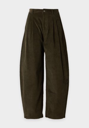 Femme aux longs cheveux raides portant un t-shirt noir et un pantalon large taille haute couleur olive foncé, debout les mains dans les poches sur un fond blanc.