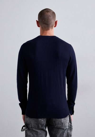 Navy blue knit sweater with long sleeves, ribbed cuffs, and a rounded neckline, worn with gray patterned trousers. Shot from the back.