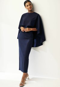 Marinblå klänning med en flödande kap design, elegant silhuett, mjuk textur och metallic blockklack-sandaler.
