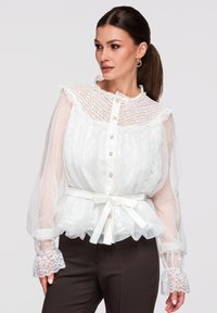 Femme portant un chemisier en dentelle blanche avec des manches bouffantes transparentes, des boutons en perles, une ceinture nouée à la taille et un pantalon foncé, regardant sur le côté.