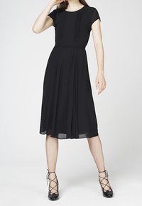 Schwarzes Kleid mit kurzen Ärmeln, plissiertem Oberteil und fließendem Rock. Aus leichter Stoffqualität, mit einem runden Ausschnitt und mehrlagigem Saum.