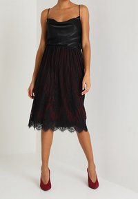 Top in satin nero con spalline sottili, che si trasforma in una gonna plissettata color bordeaux con dettagli in pizzo nero sull'orlo. Abbinata a tacchi rossi.