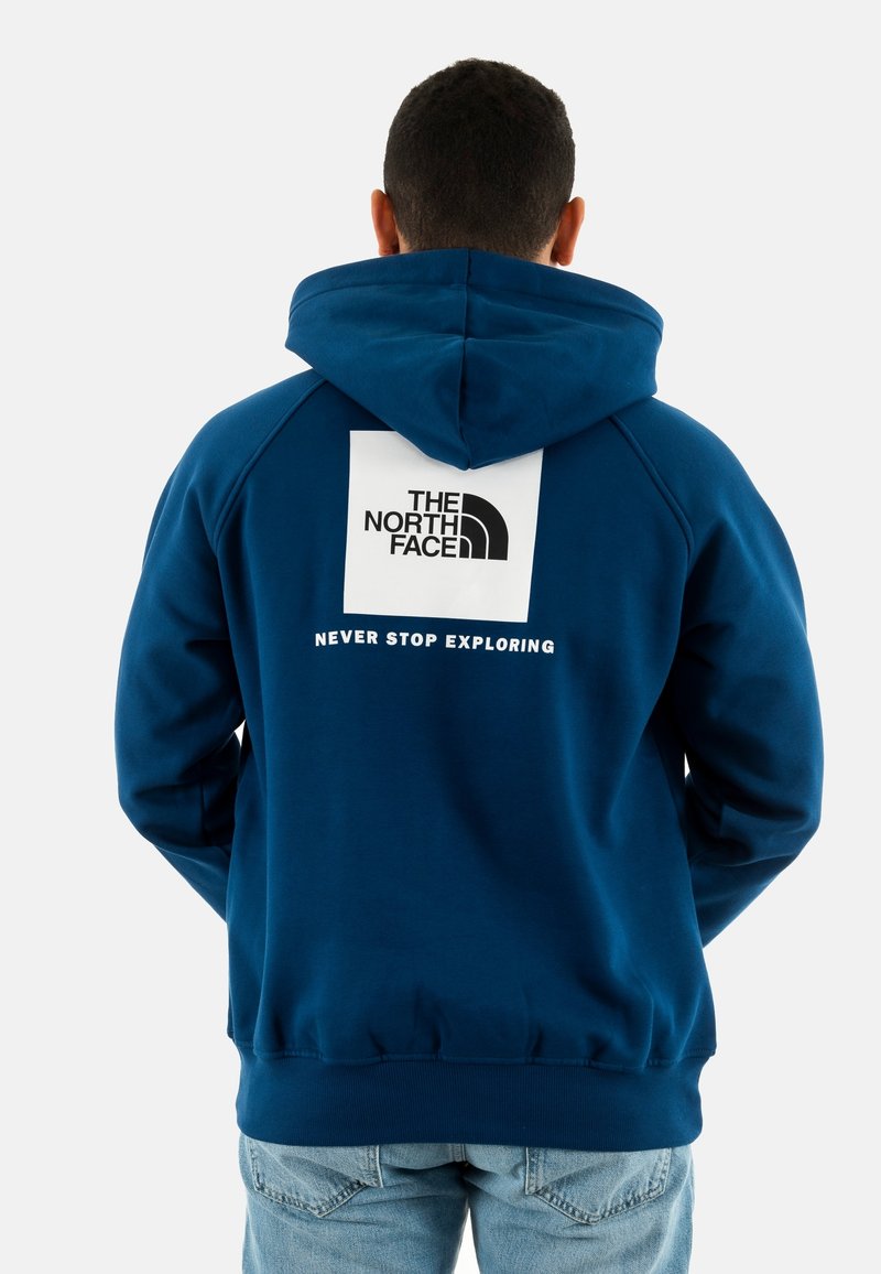 Hombre vistiendo una sudadera con capucha azul con el logo de The North Face y el texto "Nunca dejes de explorar" en la parte posterior, combinado con jeans de color azul claro.