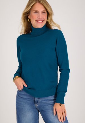 Turkoois geknoopte trui gemaakt van zachte breimateriaal, met lange mouwen en een geribbelde zoom. Draagt samen met blauwe jeans en gouden accessoires.