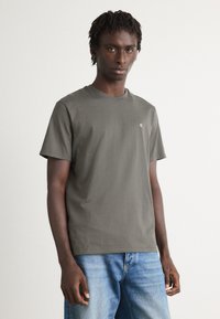Ung mand med korte dreadlocks iført en olivengrøn CK t-shirt og blå jeans står mod en ensfarvet lys baggrund.