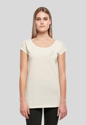 Femme aux cheveux blonds raides portant un haut beige à manches courtes avec un petit contour de cerise sur le côté gauche de la poitrine et un pantalon noir.