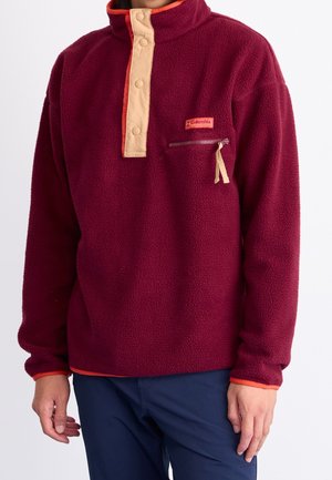 Person iført en burgunder fleece-pullover med beige trykknapper og lynlåslomme på brystet, kombineret med navyblå bukser.