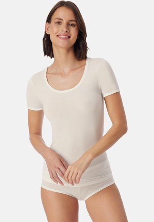 Ung kvinde med kort brunt hår, der smiler, iført en tætsiddende hvid kortærmet top med rund hals og hvide trusser, stående mod en ensfarvet baggrund.