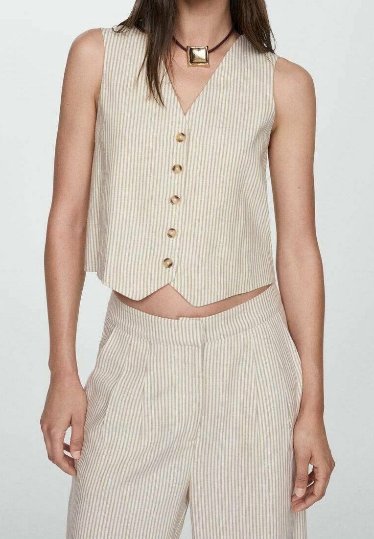 Femme portant un gilet sans manches boutonné à rayures verticales beige et blanc et un pantalon taille haute assorti avec un collier pendentif carré.