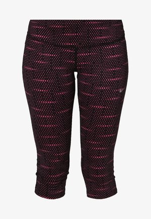 Femme portant un t-shirt de sport rose à manches courtes, des leggings capri noirs et roses à motif, et des chaussures de course noir et rose, debout les jambes croisées.