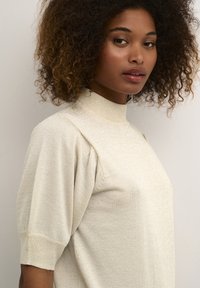 Pull léger couleur crème à manches courtes avec un col haut, texture douce et légère brillance. Présente des détails froncés aux épaules.