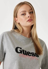 Šedé tričko s krátkým rukávem, s černým texturovaným logem "Guess" a červeným ručně psaným textem, vyrobené z bavlny.