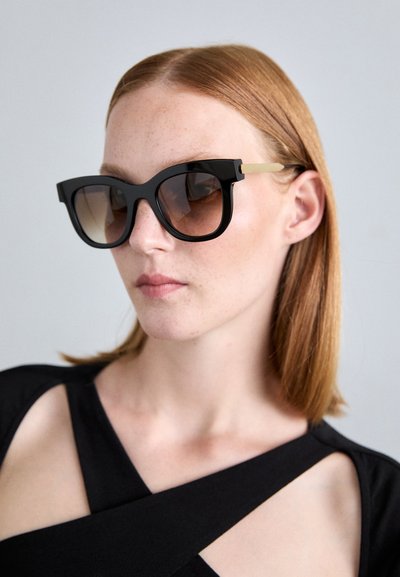 Mujer con cabello rojo liso que lleva grandes gafas de sol negras con brazos dorados y una blusa recortada negra frente a un fondo sencillo.