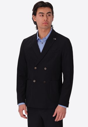 Uomo che indossa un blazer nero doppiopetto sopra una camicia azzurro chiaro, in piedi contro uno sfondo bianco uniforme.