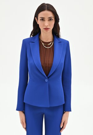 Femme portant un costume bleu roi sur mesure avec une veste à un bouton sur un haut marron plissé et un collier doré à plusieurs rangs, debout devant un fond blanc.