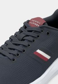 Baskets gris foncé avec dessus en tissu, lacets noirs, écusson logo blanc et rouge sur le côté, et étiquette rouge sur la languette avec l'inscription « Tommy Hilfiger ».