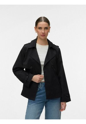 Femme portant une veste noire à double boutonnage sur un haut blanc et un jean bleu, debout devant un fond blanc uni, faisant face à l'avant.