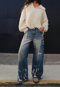 Personne portant un pull en maille crème oversize et un jean large en denim délavé, debout les mains dans les poches sur le trottoir.
