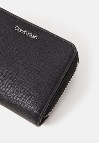 Black textured leather wallet with silver hardware, featuring a prominent Calvin Klein logo and a zippered closure along the edge.