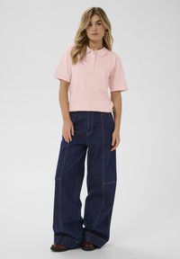 Polo rose clair avec un col chemise et des manches courtes, assorti à un jean en denim bleu foncé à jambes larges avec des surpiqûres contrastantes.