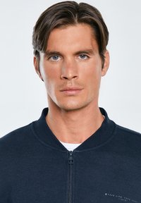 Giacca zippata blu navy con tessuto texturizzato, colletto a coste e un sottile dettaglio del logo sul petto. Indossata sopra una camicia bianca.
